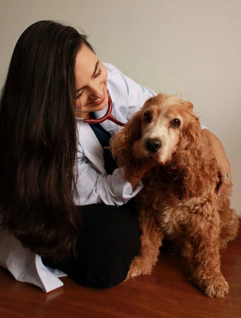Dra. Marcella Moreira de jaleco e estetoscópio, sorrindo ao lado de um cachorro cocker